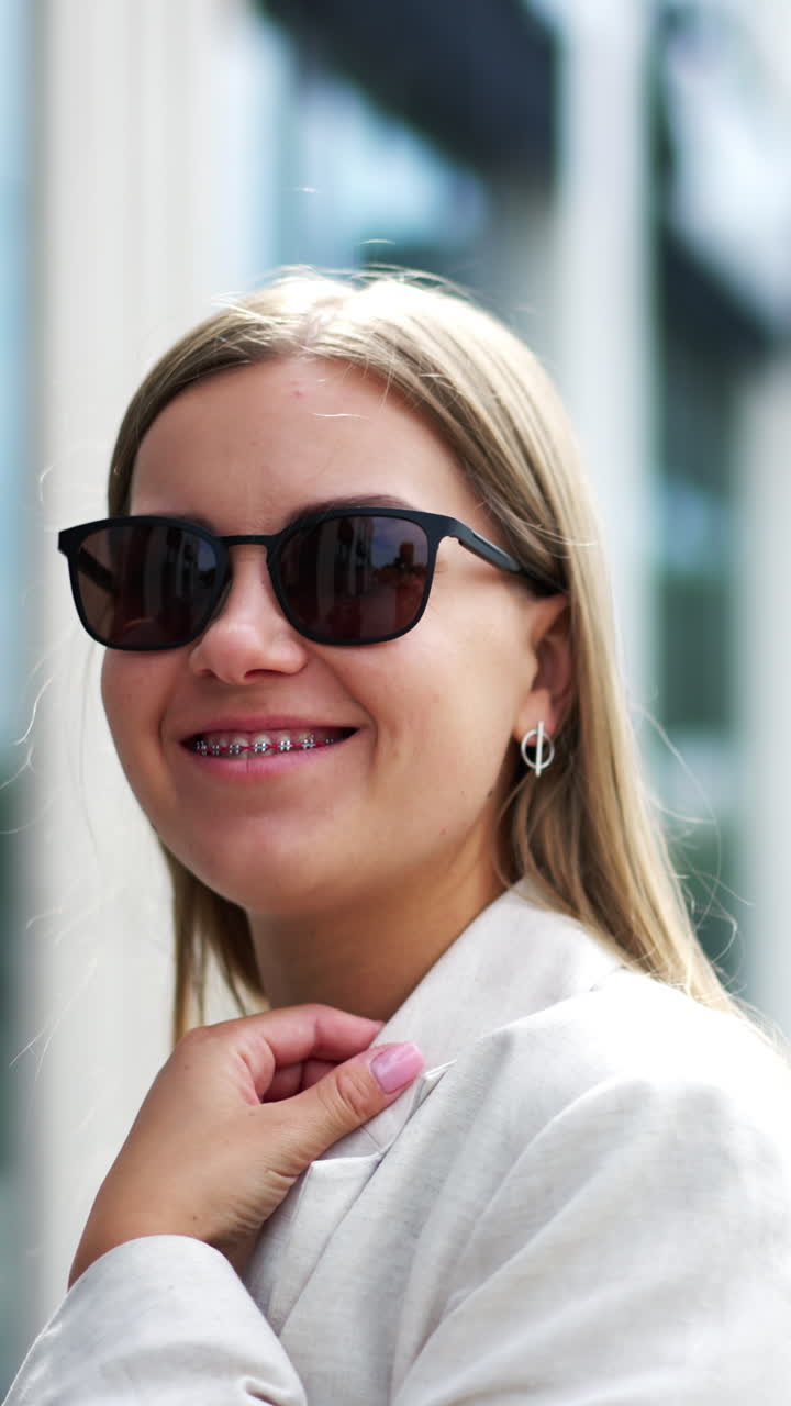 Blonde woman wearing black sunglasses standing in the street. Smiling lady in white jacket and black top posing to the camera. Blurred backdrop. Vertical video