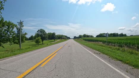 pov - driving on a rural county road past trees and lush, green fields in late summer in central iowa