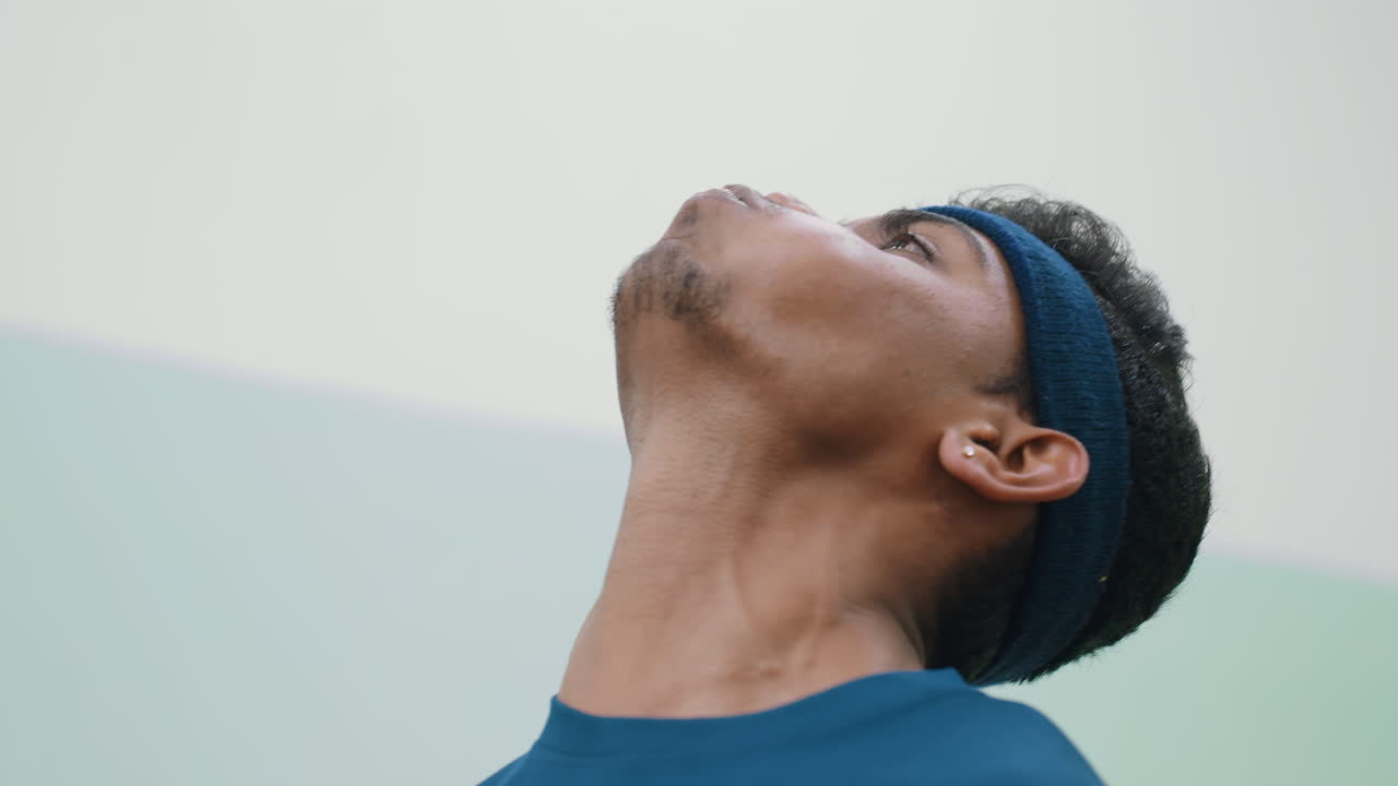 Young man in blue headband tilting head back and blowing upward to keep tennis ball in air, smiling and laughing during playful challenge, captured in close side angle showing joy