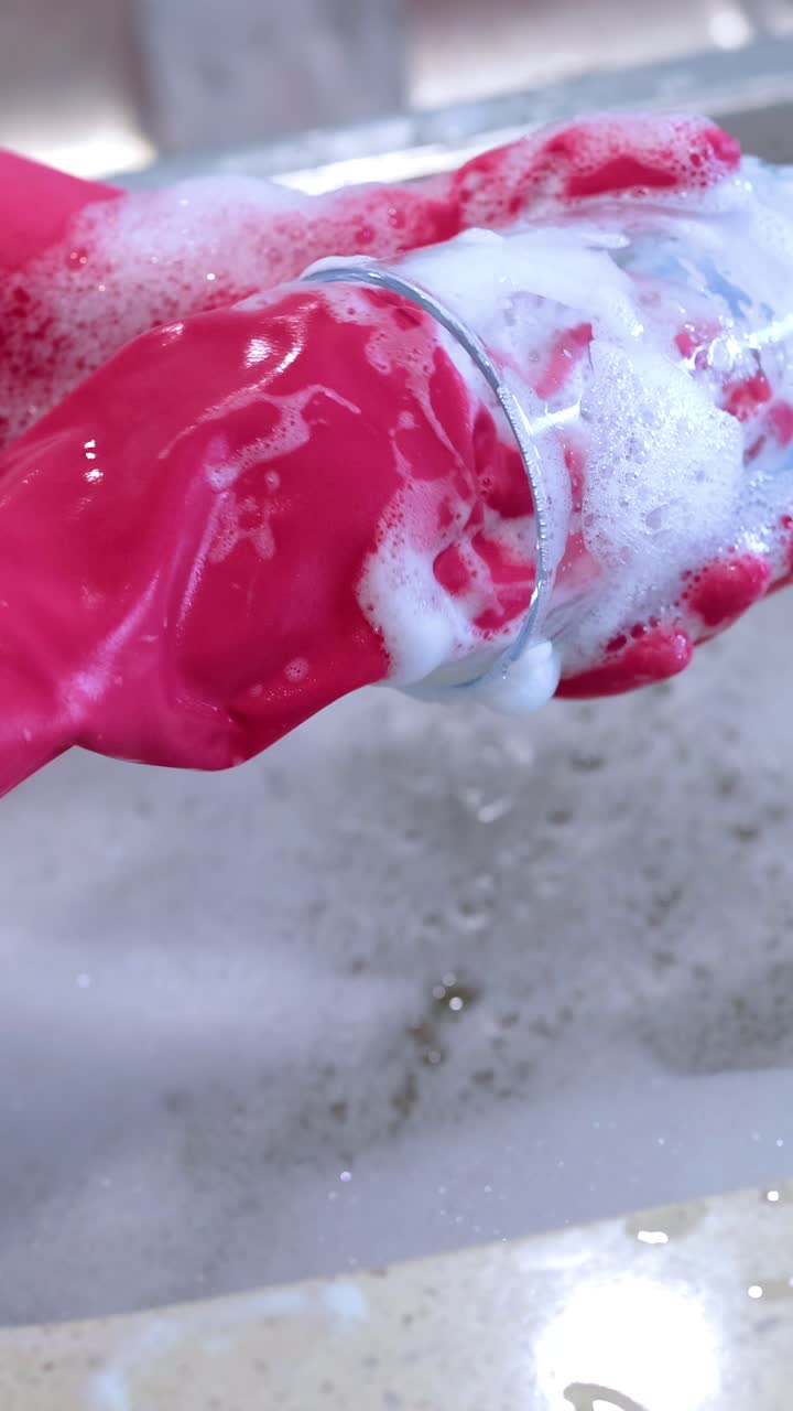 Mani con guanti rosa lavano un bicchiere con acqua saponata nel lavello della cucina, sotto una luce intensa.