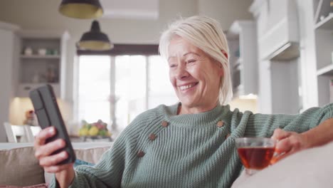happy senior caucasian woman sitting on sofa in living room, using smartphone and drinking tea