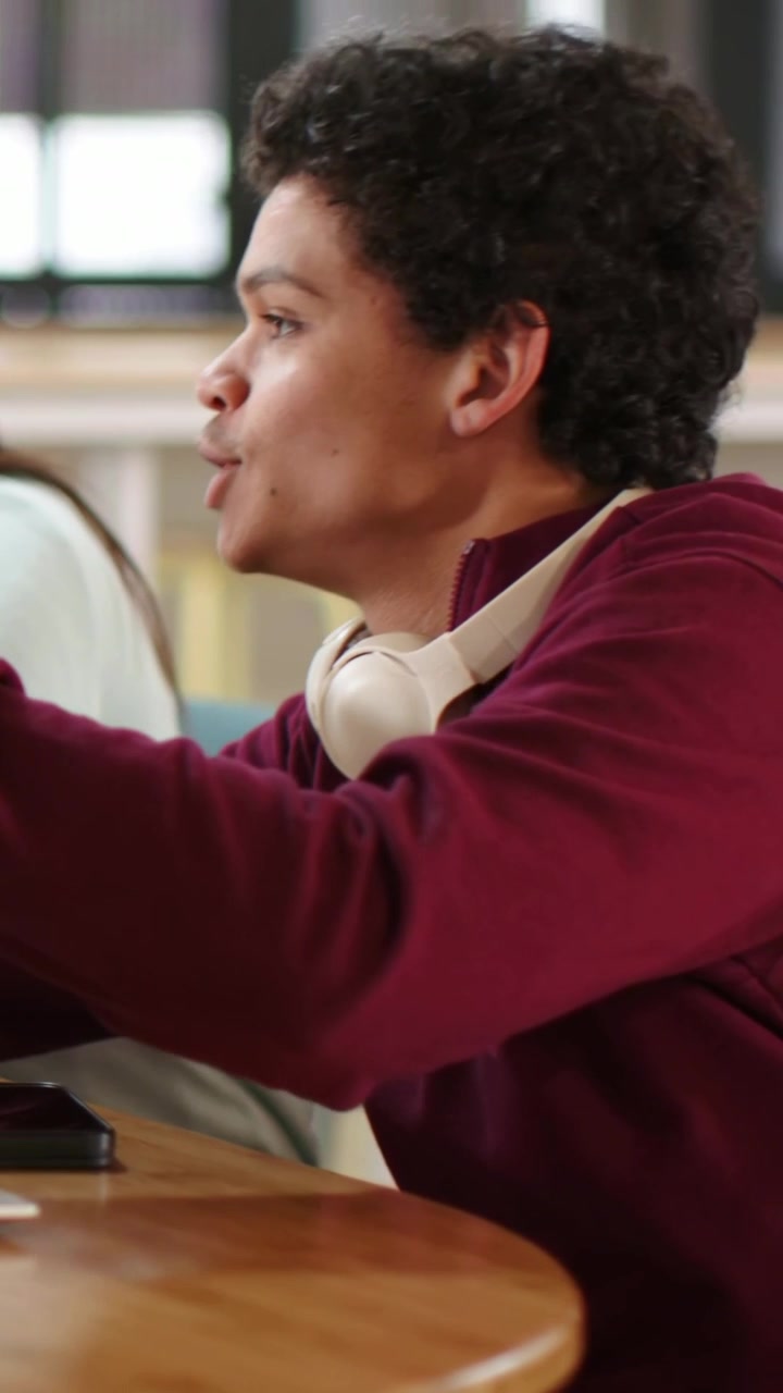 Vertical video: Reaching student in maroon top grabbing notebook and explaining to peer at library