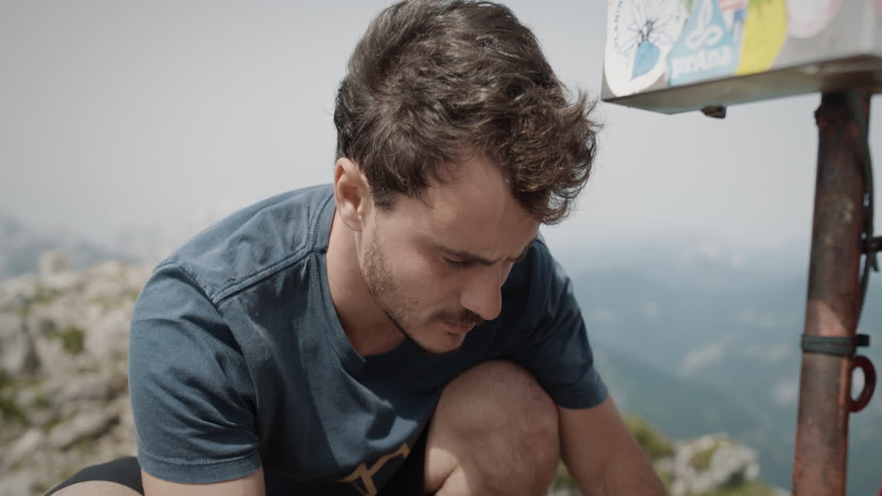 joven excursionista en cuclillas en la cima de la montaña junto a una caja para sellar su folleto de senderismo