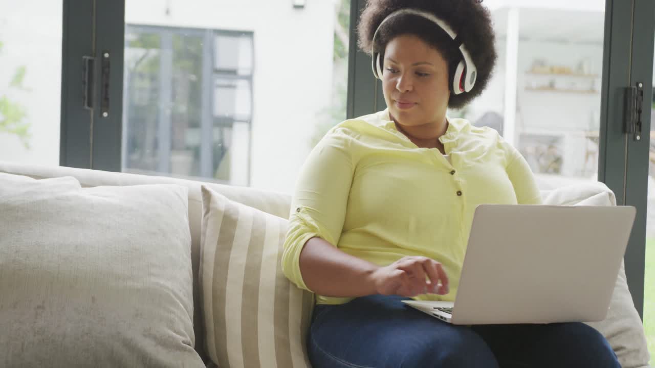 Video of happy plus size african american woman with headphones sitting on sofa with laptop