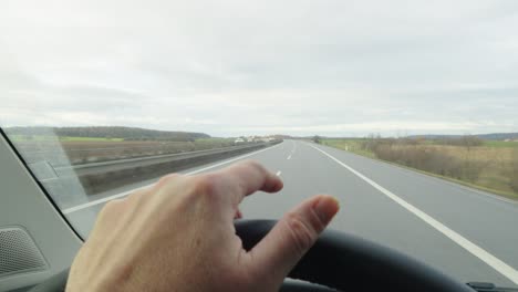 Punto-De-Vista-De-Una-Mano-En-El-Volante-Golpeando-El-Ritmo-Mientras-Conduce-Por-La-Carretera