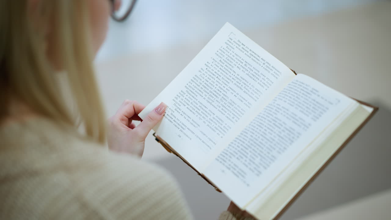 책의 다음 페이지로 전환하는 은 손을 가진 여자의 가까운 후면 뷰, 잘 조명된 환경에서 볼 수 있는 안경의 부분적인 흐릿함, 텍스트에 부드러운 초점 및 섬세한 손