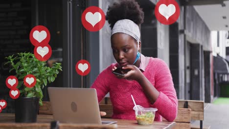 animación de los iconos del corazón de las redes sociales sobre una mujer afroamericana con computadora portátil y teléfono inteligente en un café