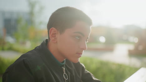 Close up of young boy outdoors lifting head thoughtfully with soft sunlight illuminating face, capturing quiet moment of reflection and focus in natural environment with blurred green background