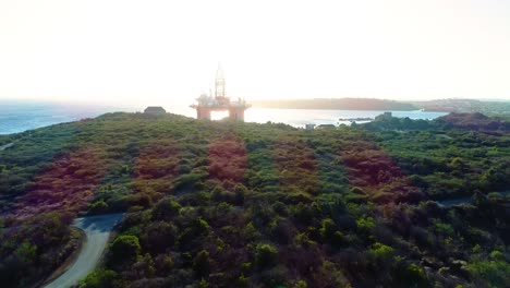 Off-shore-oil-rig-silhouette-at-edge-of-curacao-backlit-at-sunset,-drone-dolly