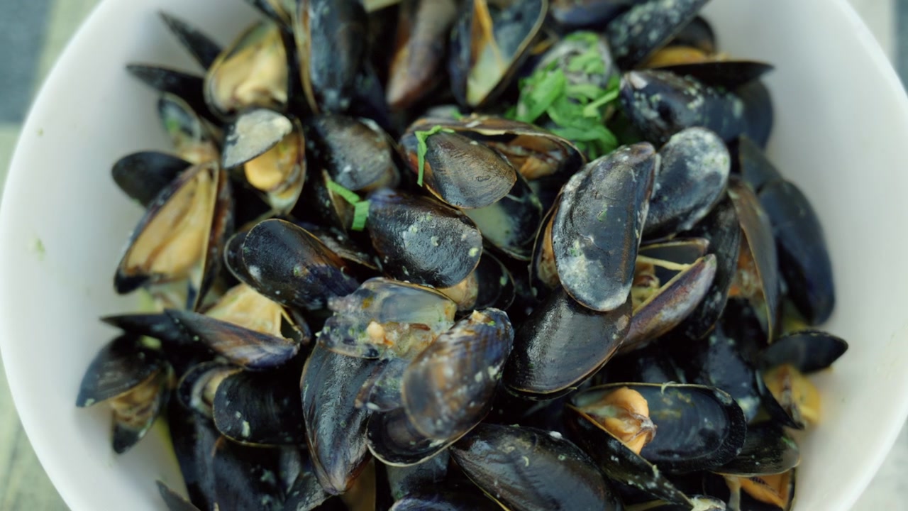 Mejillones recién cocinados servidos en un cuenco blanco, cubiertos con una salsa cremosa y adornados con hierbas verdes. Comida de mariscos.