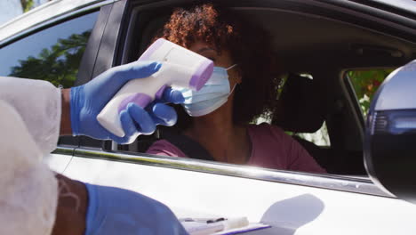 African-american-woman-with-face-mask-sitting-in-car-having-temperature-measured-by-medical-worker