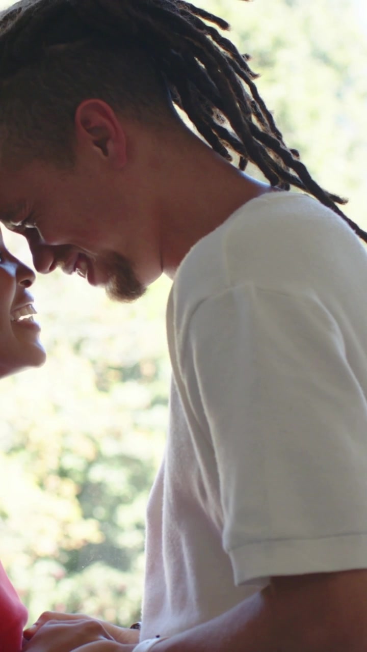 Vertical video: Smiling man holding hands by window, leaning to her in red top, wearing white tee