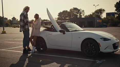 Happy-couple,-a-blond-guy-with-glasses-in-a-checkered-shirt-and-his-blonde-girlfriend-in-a-white-sweatshirt-stand-near-a-white-convertible-holding-hands-and-looking-at-each-other-on-a-large-square-in-the-city-in-the-morning