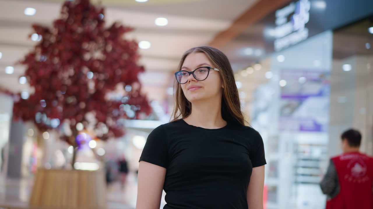 mujer joven con gafas y una camiseta negra caminando con confianza a través de un centro comercial moderno, atmósfera elegante con tiendas borrosas, luces brillantes y compradores que pasan en el fondo