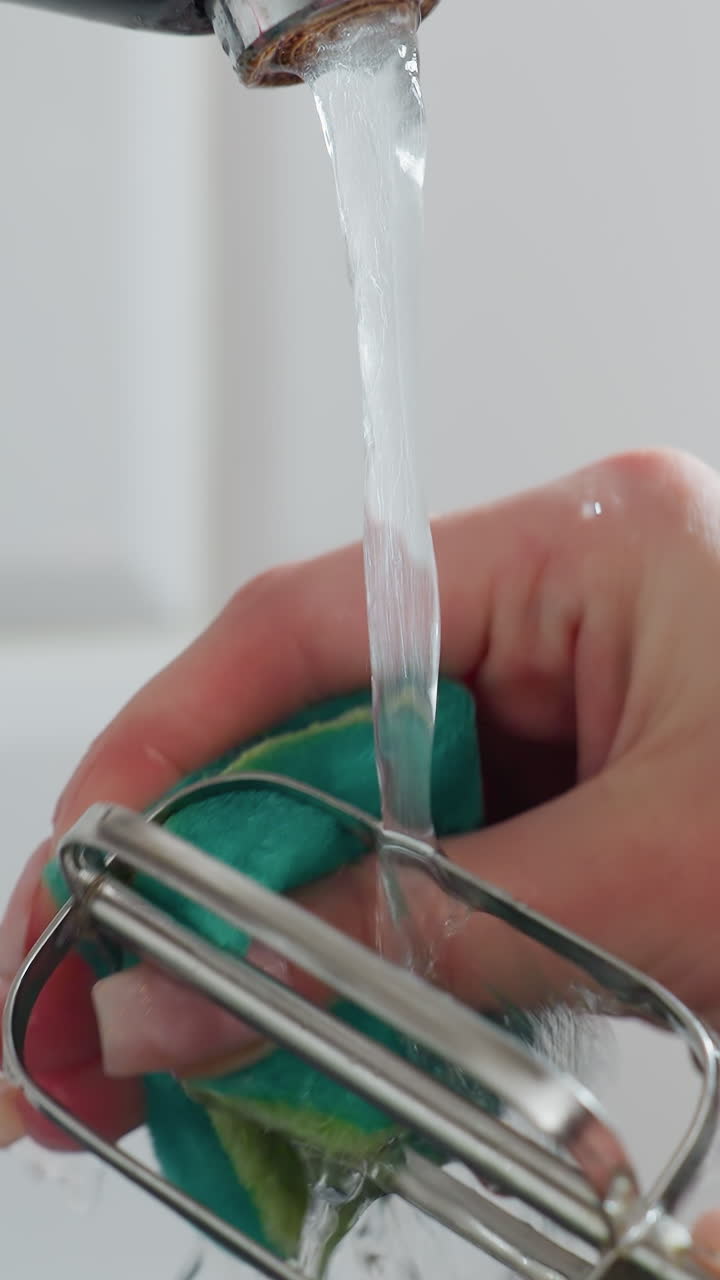 primer plano de un grifo en funcionamiento con una persona lavando el batedor usando una esponja verde bajo el agua que fluye en la cocina, salpicaduras de agua alrededor del batedor mientras se limpia, configuración moderna de la cocina