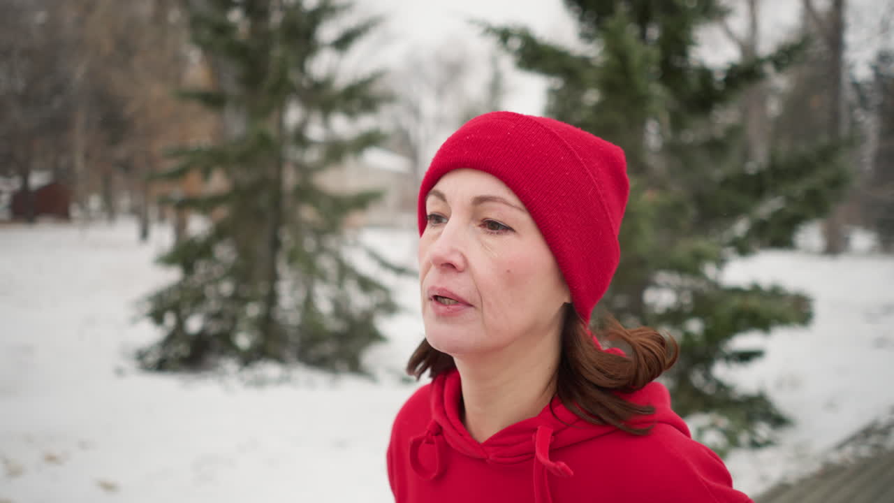 mujer de mediana edad con gorra roja y sudadera con capucha corriendo al aire libre durante el invierno, respirando pesadamente con una atmósfera serena y brumosa, rodeada de árboles cubiertos de nieve, pinos de hoja perenne