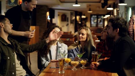 handsome male waiter is coming to group of friends sitting at table and taking order. young people are talking and laughing, waiter is very friendly and helpful.