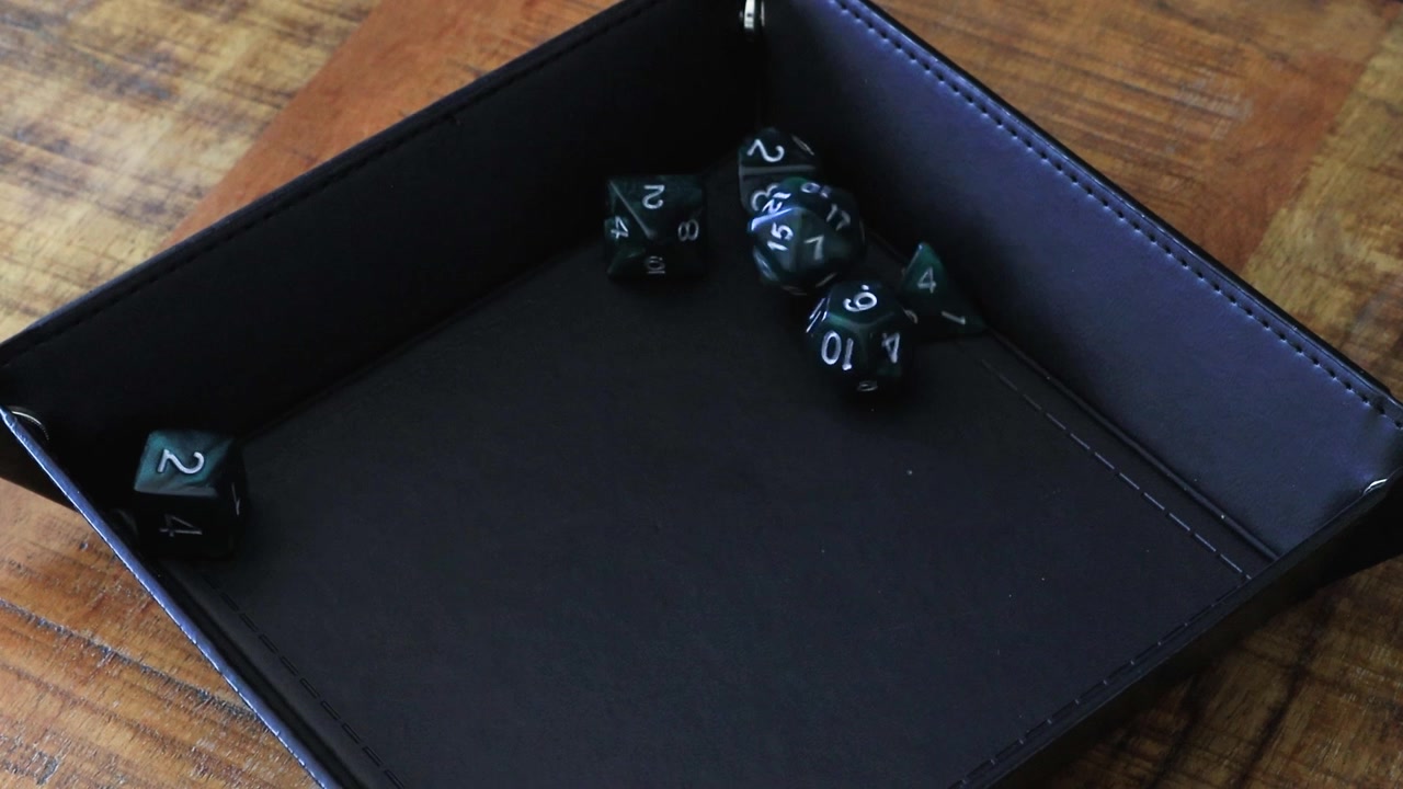 Un giocatore lancia un D20 in un vassoio portadadi nero che si trova su un tavolo di legno. All'interno del vassoio portadadi ci sono già alcuni dadi come un D12, un D6, un D10 e un D4.