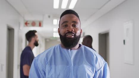 Retrato-En-Vídeo-De-Un-Trabajador-Médico-Afroamericano-Sonriente-Y-Barbudo-Vestido-Con-Bata-Quirúrgica-En-El-Pasillo