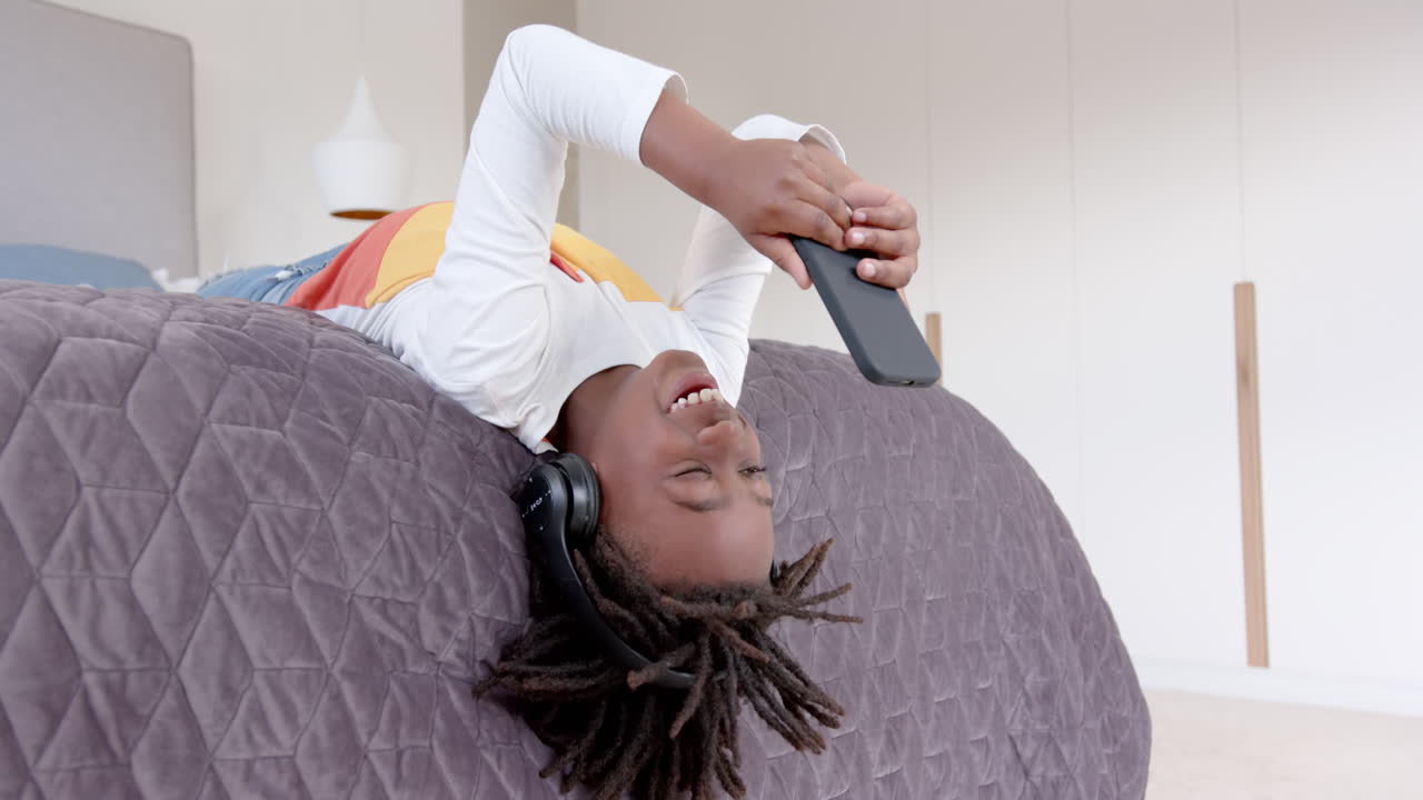 un niño afroamericano feliz con auriculares acostado en la cama usando un teléfono inteligente en casa, en cámara lenta