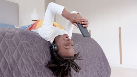 un niño afroamericano feliz con auriculares acostado en la cama usando un teléfono inteligente en casa, en cámara lenta