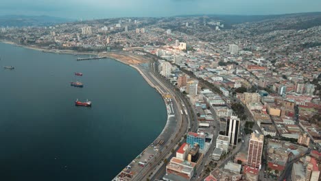 Aerial-dolly-in-of-Valparaiso-hillside-city-buildings-and-Sea-Port-with-container-cargo-ships-sailing-near-the-coast,-Chile