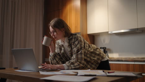 a young beautiful business woman works from home with a cup of coffee and looks into a laptop screen standing at the table. view mail and drink coffee. remote work from home