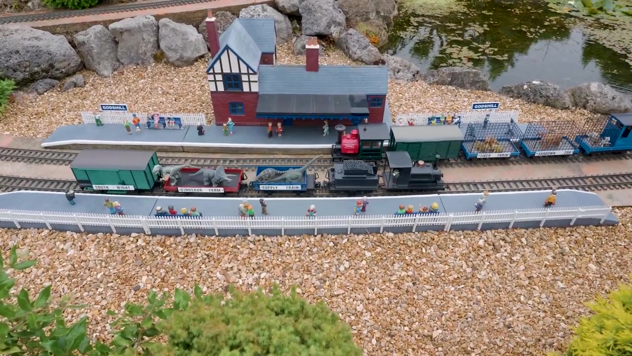 Modellino di treno che lascia la stazione di Godshill nel villaggio modello di Godshill, mentre la telecamera segue la locomotiva in miniatura che si allontana oltre la banchina, piccole figure e l'edificio della stazione in un'esposizione all'aperto.