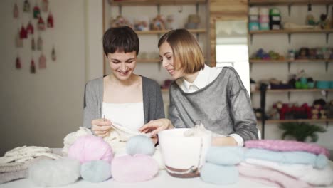 Two-woman-friends-study-knitting-yarn-wool-sitting-on-table-in-home-room