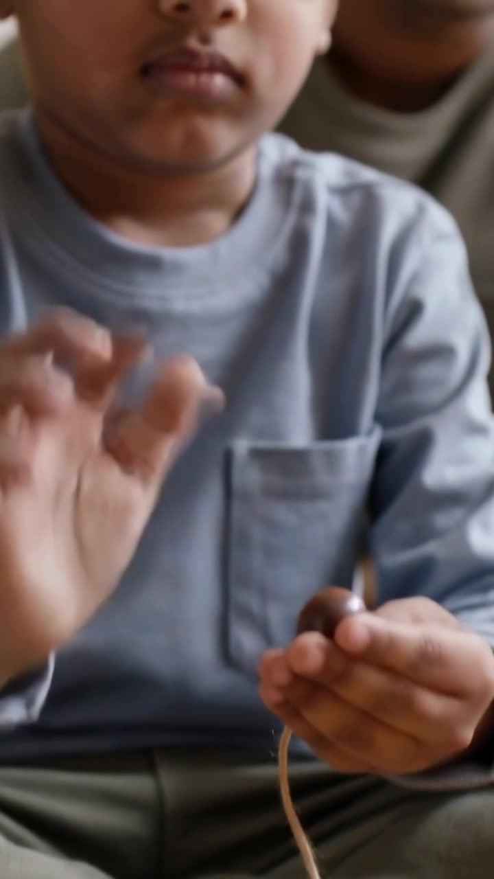 Vertical video: Picking bead child in grey shirt threading beads on twine at home rug, dad teaching