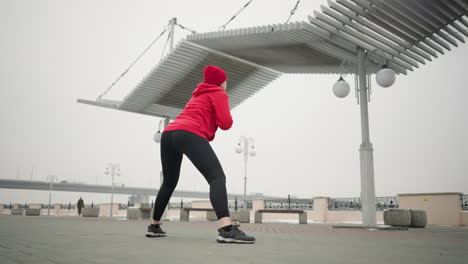 woman in red hoodie performing stretching workout outdoors under modern urban canopy structure, with someone walking in blurred background and a distant bridge with cars visible in foggy weather