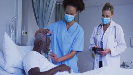 Diverse-female-doctors-putting-oxygen-mask-on-male-patient-in-hospital-bed-all-wearing-face-masks