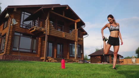 una mujer delgada y hermosa en ropa deportiva con una prensa abierta se está preparando para comenzar a entrenar en el césped cerca de su casa flexionando sus piernas, rodillas, espinillas y muslos con ejercicios de calentamiento