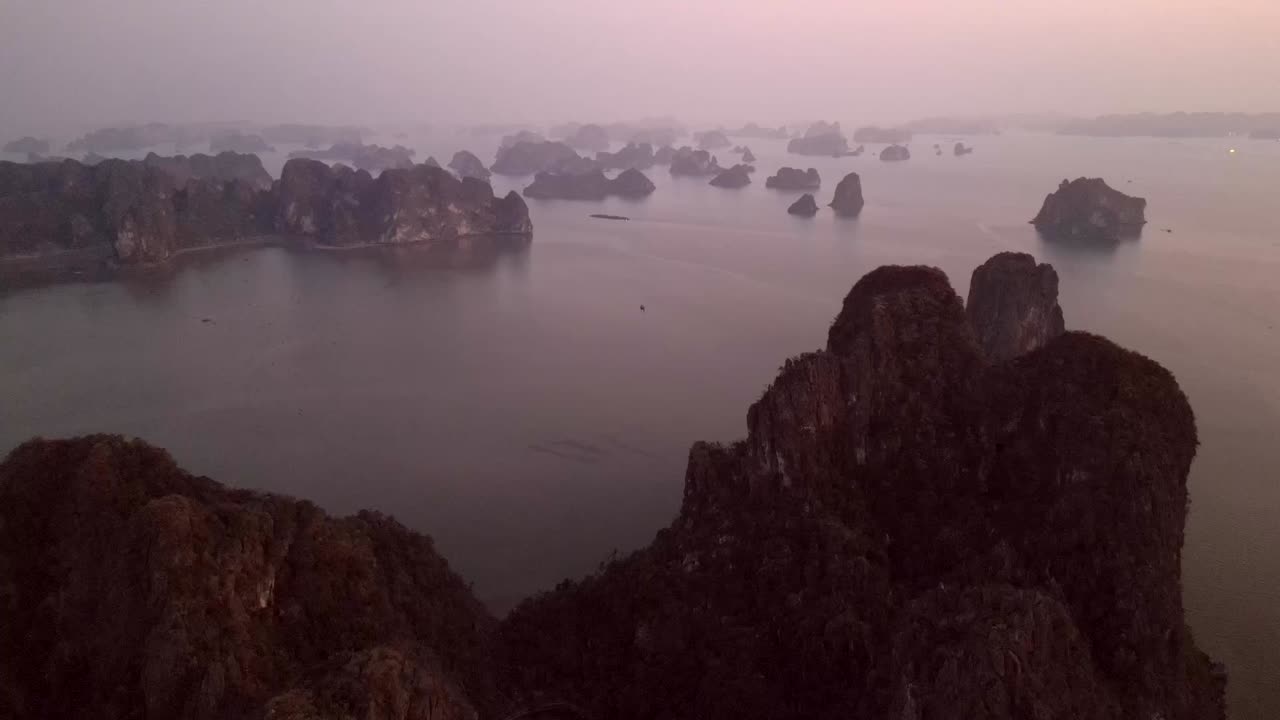 Drone flies over towering limestone karsts scattered across a calm bay during the blue hour, capturing layers of rocky islets fading into the mist.