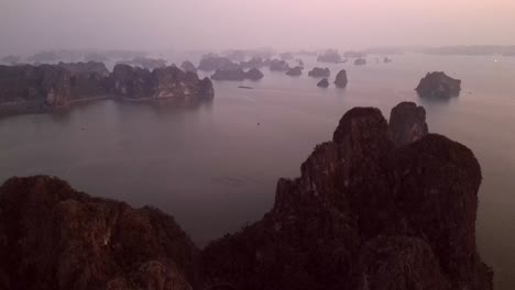 drone flies over towering limestone karsts scattered across a calm bay during the blue hour, capturing layers of rocky islets fading into the mist.