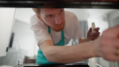 view from inside the oven a confident guy a blond cleaner in a white t-shirt and a blue apron washes the walls of the oven using a rag and a cleaning product in a modern apartment in the kitchen