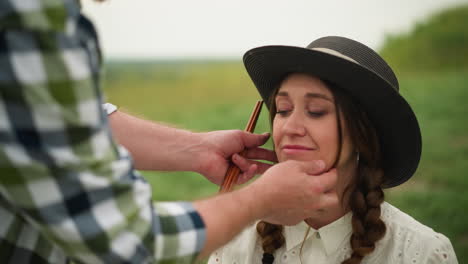 a craftsperson in a checked shirt gently adjusts the hat of a woman in a white dress, her braided hair and smile brightening the serene green field setting