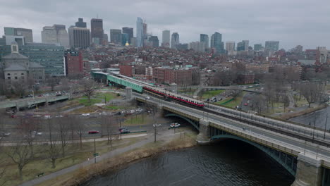 Imágenes-Aéreas-De-La-Unidad-De-Metro-Cruzando-El-Río-Sobre-El-Puente-Y-Deteniéndose-En-La-Estación.-Paisaje-Urbano-En-Un-Día-Nublado-En-El-Fondo.-Boston,-Estados-Unidos