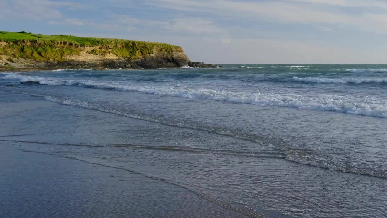 Air pasang dan ombak yang menghantam di pantai berpasir dengan tebing di kejauhan.