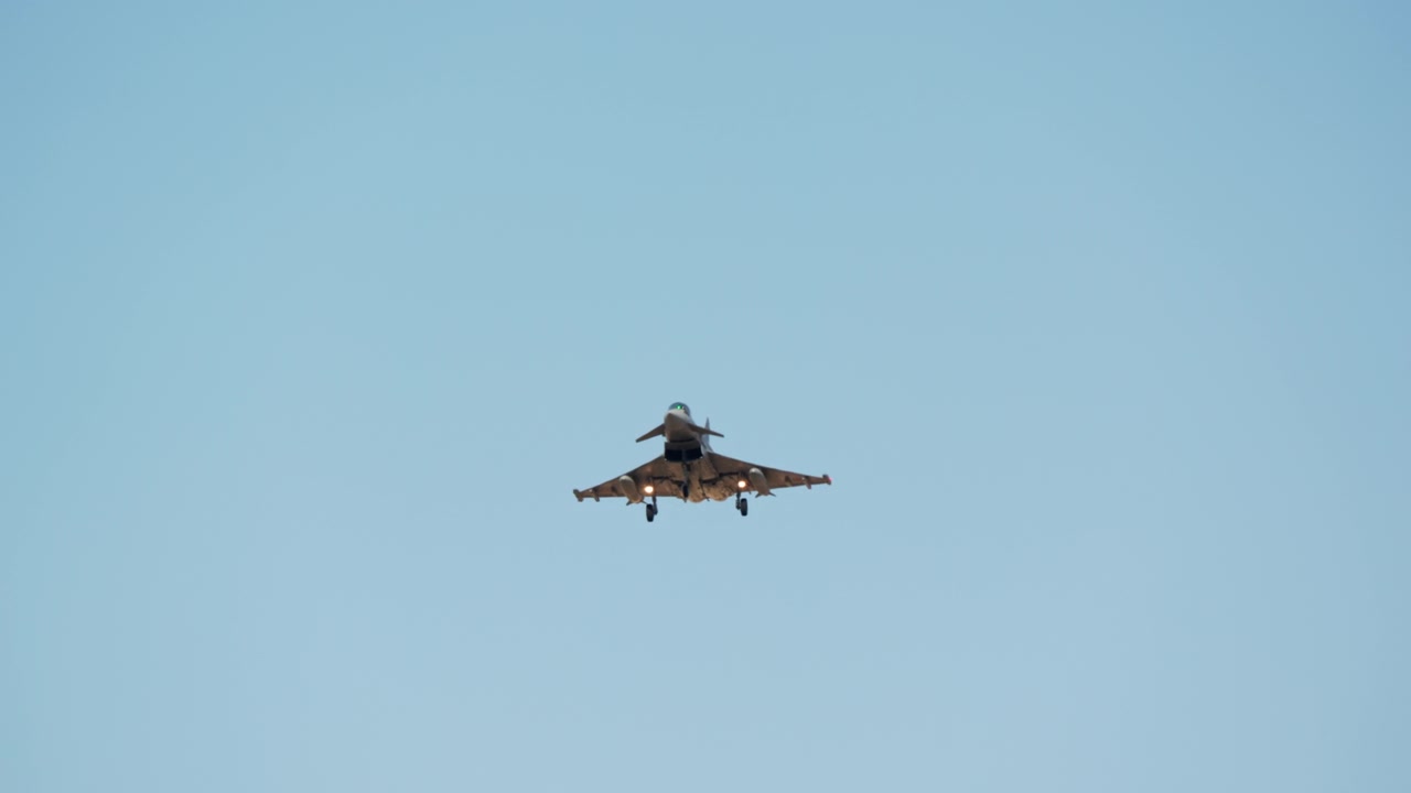 A military fighter jet with its landing gear down slowly approaches for a landing against a clear blue sky, captured in slow motion
