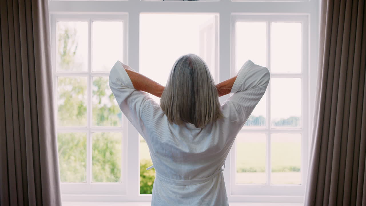 Senior Woman Opens Curtains And Stretches In Front Of Window
