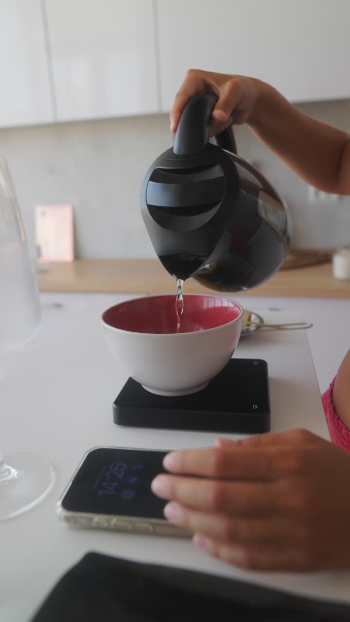 Person pouring water from a kettle into a bowl on a kitchen scale while using a smartphone