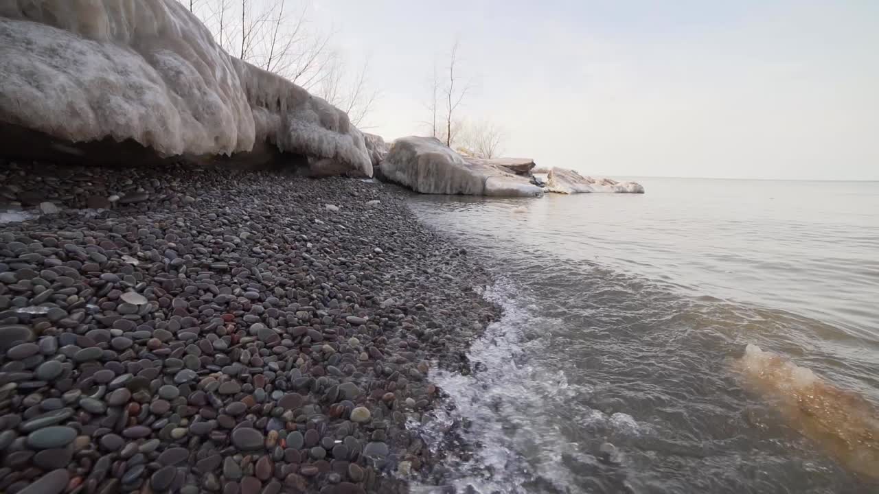 небольшие волны, медленно набегающие на галечный пляж, окруженный тающими ледяными образованиями, с маленькими кусочками льда, плавающими в холодной воде озера