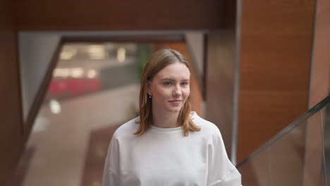 young woman riding moving escalator with hand resting on rail, smiling while looking ahead, dressed in casual white shirt, in modern shopping mall with glass railing and blurred background