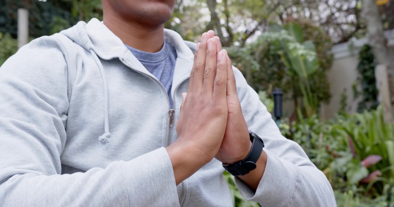 Homem africano começando com as mãos afastadas, unindo-as em busca de calma em um jardim, usando moletom e relógio.
