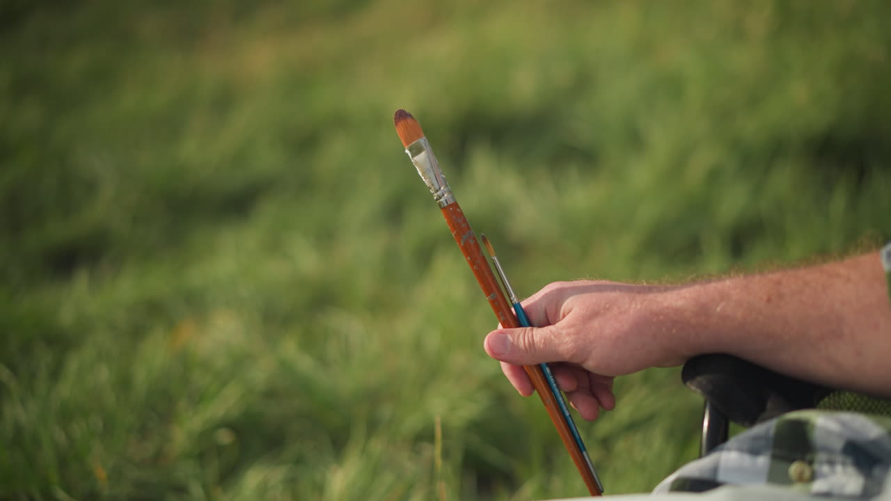 una toma de primer plano de la mano de un hombre sosteniendo dos pinceles, uno pequeño y otro muy pequeño, contra un fondo verde borroso, la escena captura la simplicidad y el enfoque en las herramientas de la creación artística.