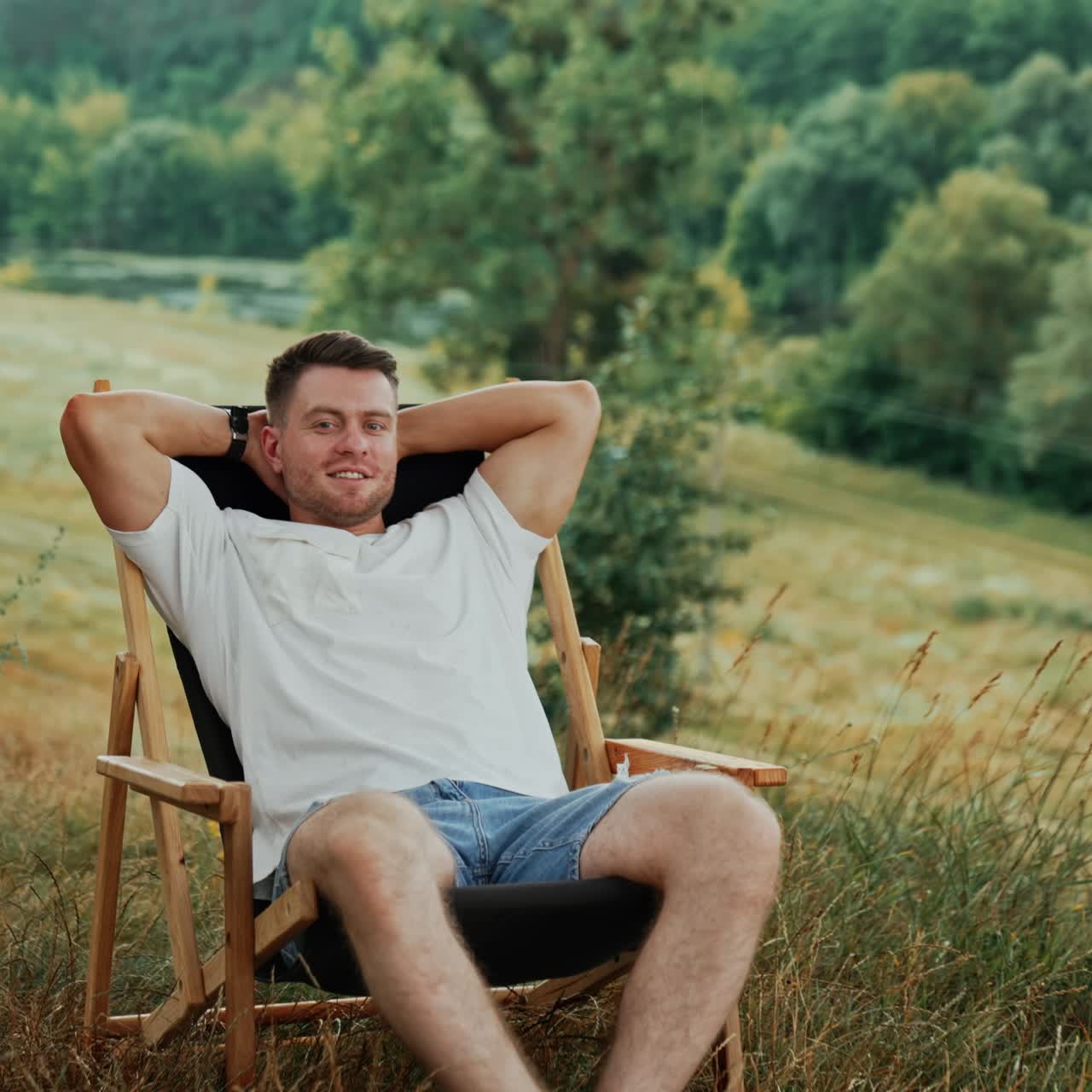 Dreamy Caucasian man sits in a chair his hands behind head. Relaxed man enjoys beautiful nature around him