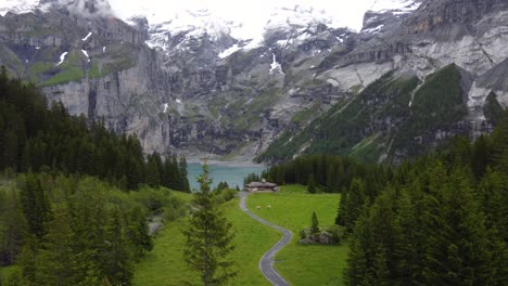 aerial descend view isolated wooden cabin chalet farm alone on green alpine meadow surrounded by alp mountains, pine trees overlook turquoise azure glacier lake oeschinensee in kandersteg, switzerland