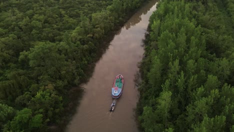 cinematic drone approaching shot of ship carrying small boat on amazon river surrounded by green rainforest trees during sunset