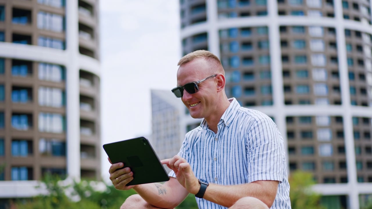 Man using tablet outdoors. Caucasian male wears sunglasses and striped shirt sitting near modern buildings.